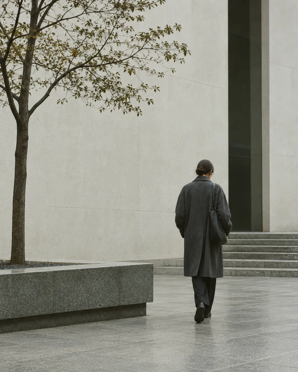 A person in a long dark grey coat walks away across a minimalist stone plaza beside a slender tree.