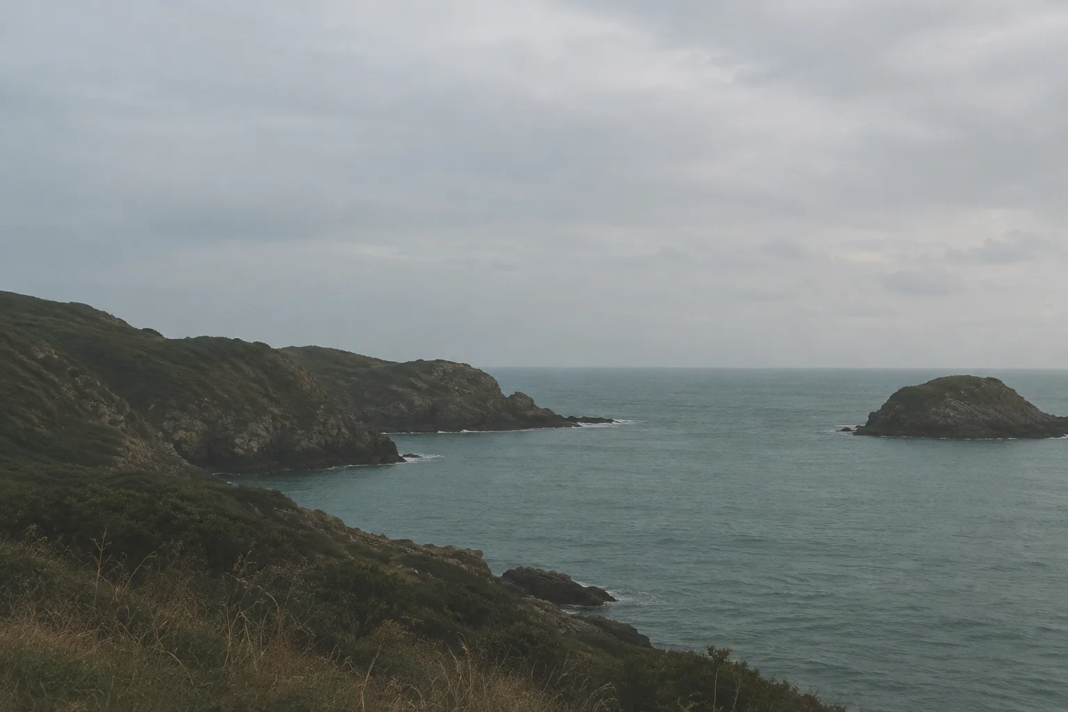 Rugged grassy cliffs jut into a calm teal sea beneath a heavy overcast sky beside a solitary rocky islet.
