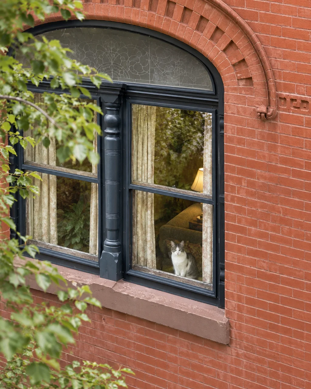 A domestic cat sits on an indoor windowsill looking out through an ornate arched window framed by red brick.