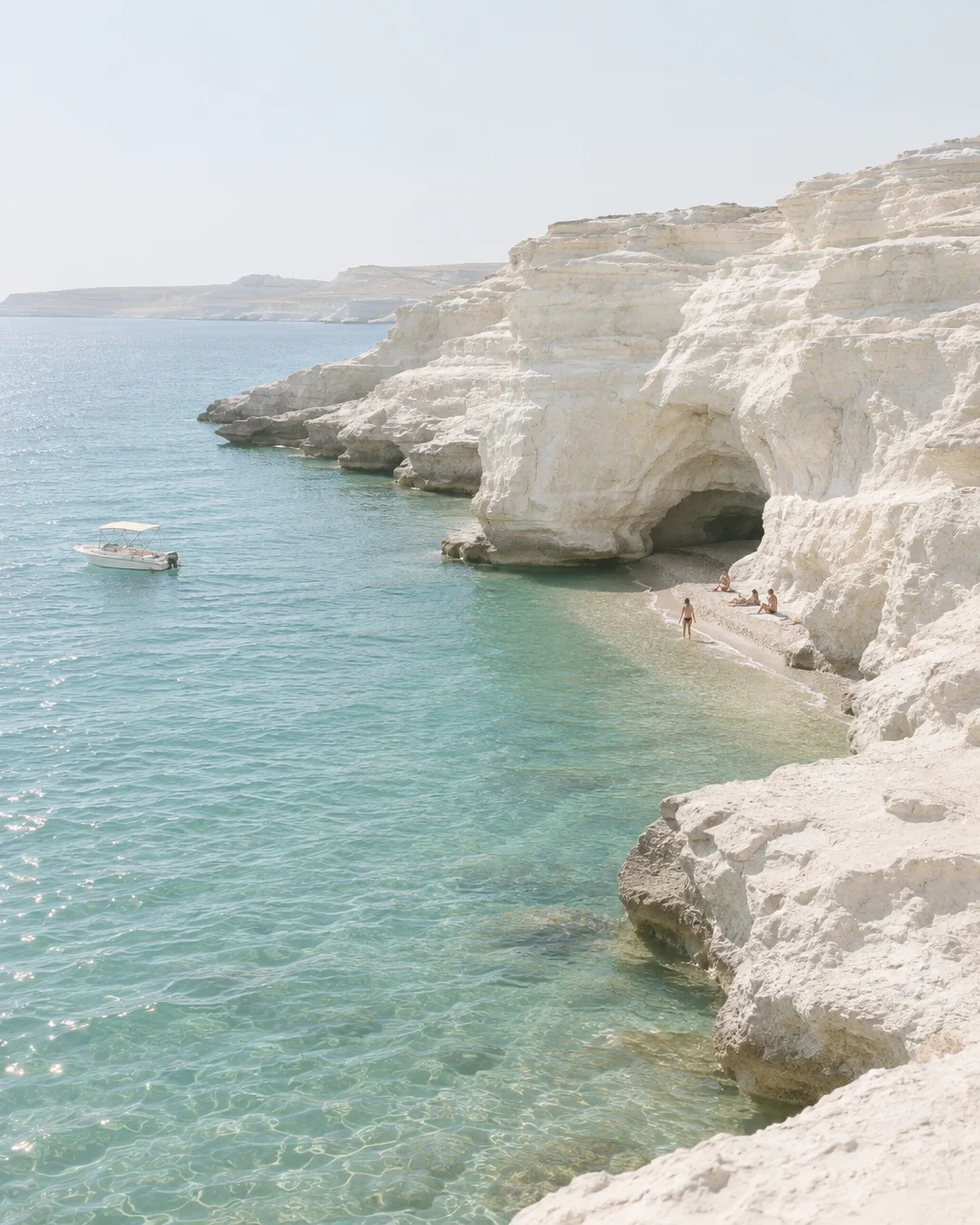 Sunbathers rest on a secluded sandy cove beneath towering white limestone cliffs beside calm turquoise water and an anchored boat.