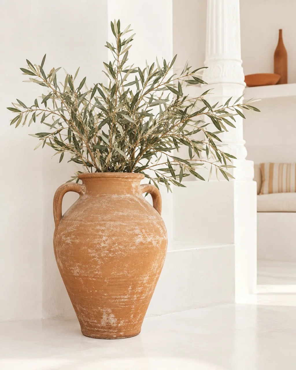 A large weathered terracotta urn filled with leafy olive branches stands inside a bright, whitewashed Mediterranean interior.