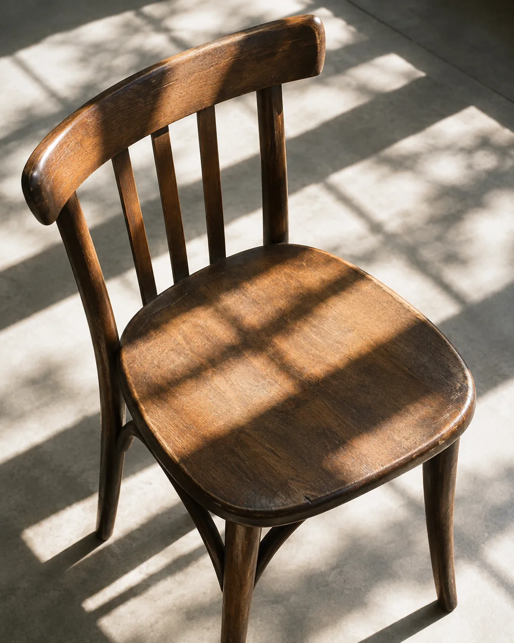 A vintage wooden chair rests on a concrete floor under geometric window shadows and soft natural light.