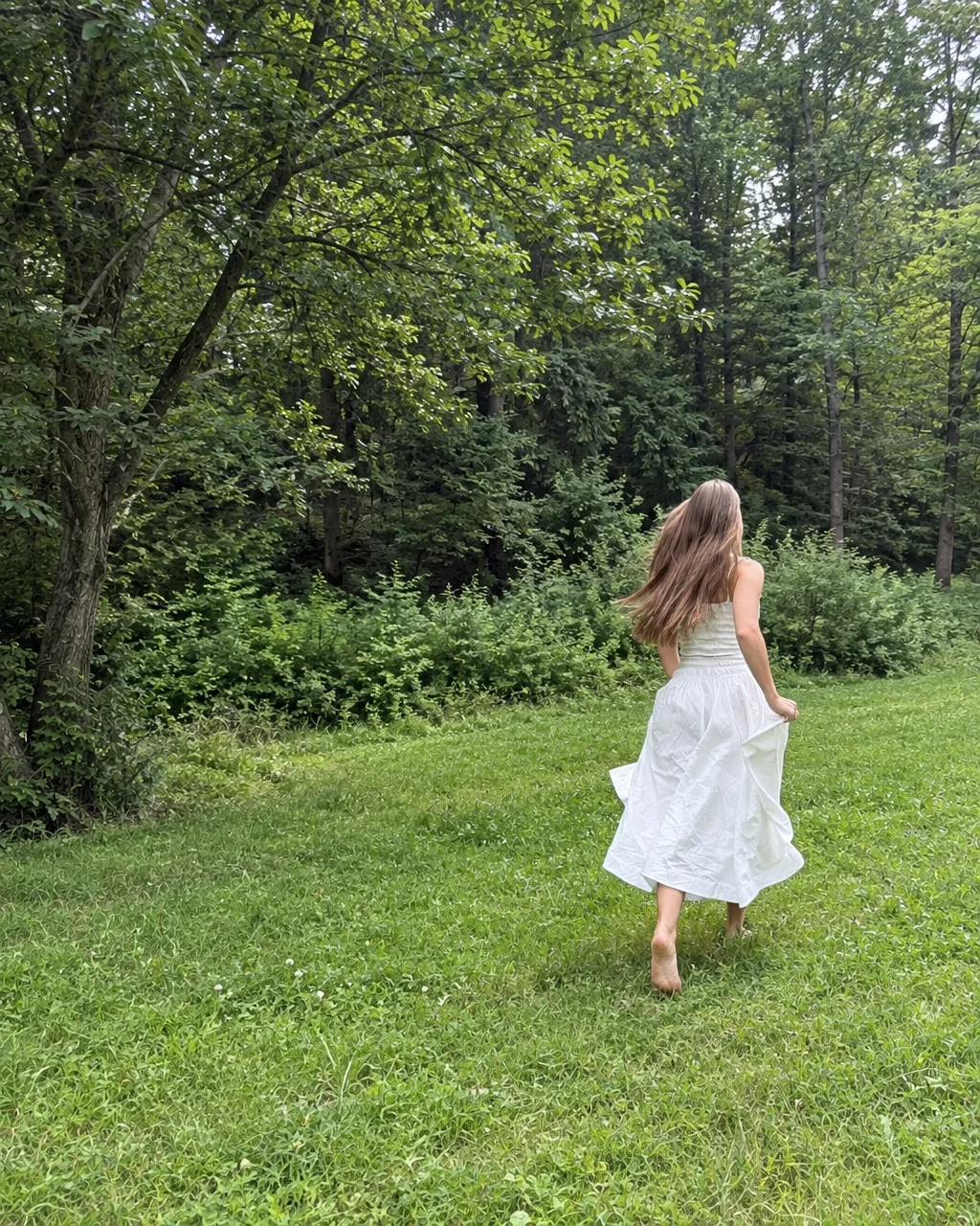 A woman in a flowing white dress runs barefoot across a grassy field toward a dense green forest.