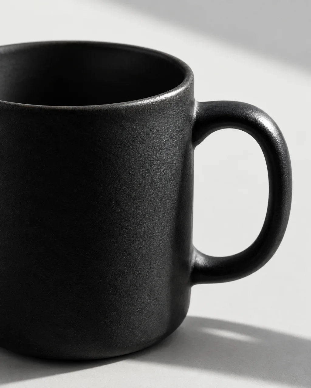 A close-up of a matte black stoneware mug casting a soft diagonal shadow on a pale neutral surface.