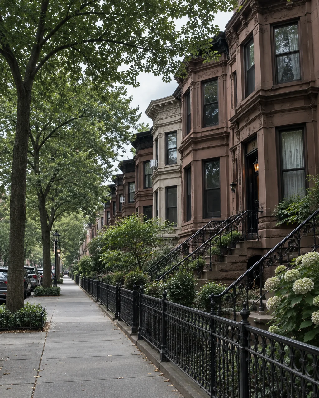 A quiet urban street lined with classic brownstone townhouses, ornate iron railings, and mature green trees.