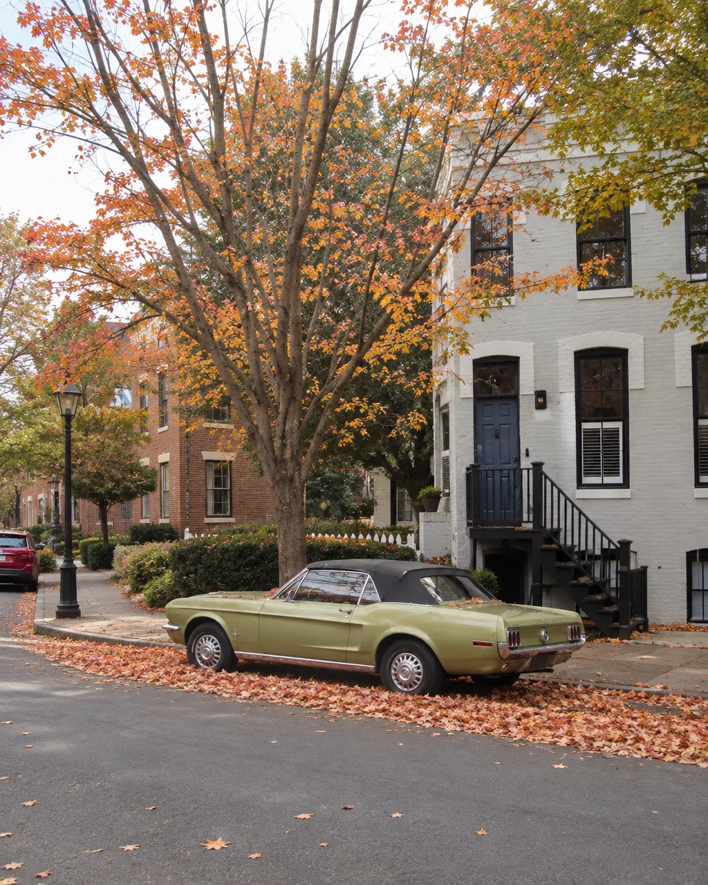 A vintage green convertible parked beside a brick townhouse, flanked by a street covered in fallen autumn leaves.