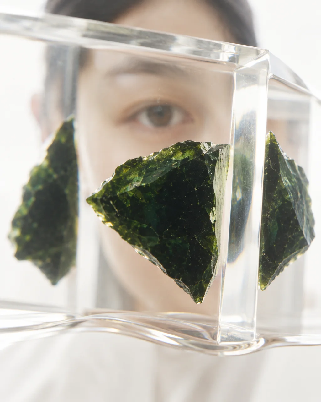 A dark green mineral fragment captured through clear refractive acrylic, with a person's soft gaze visible in the background.