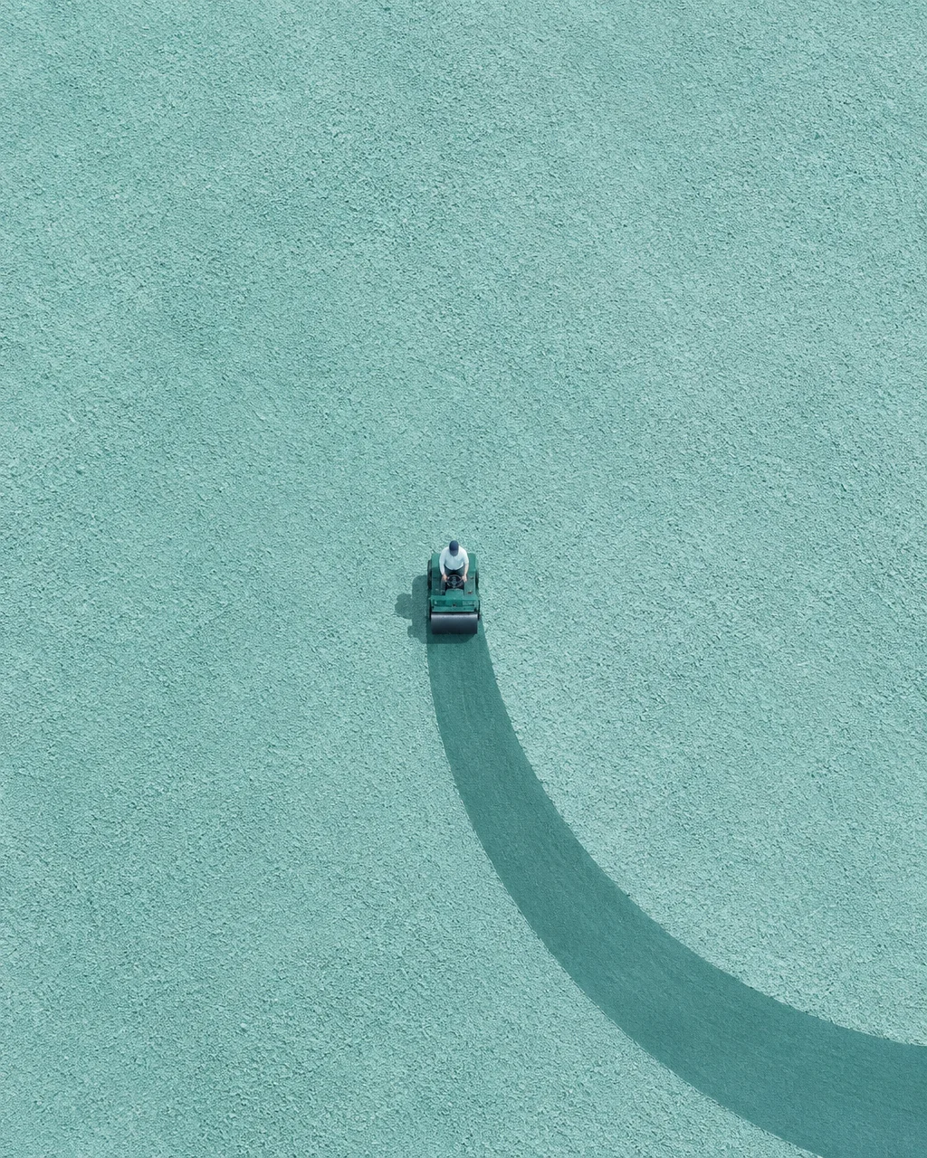 An overhead view of a small roller smoothing a sweeping curved trail across a vast textured turquoise ground.