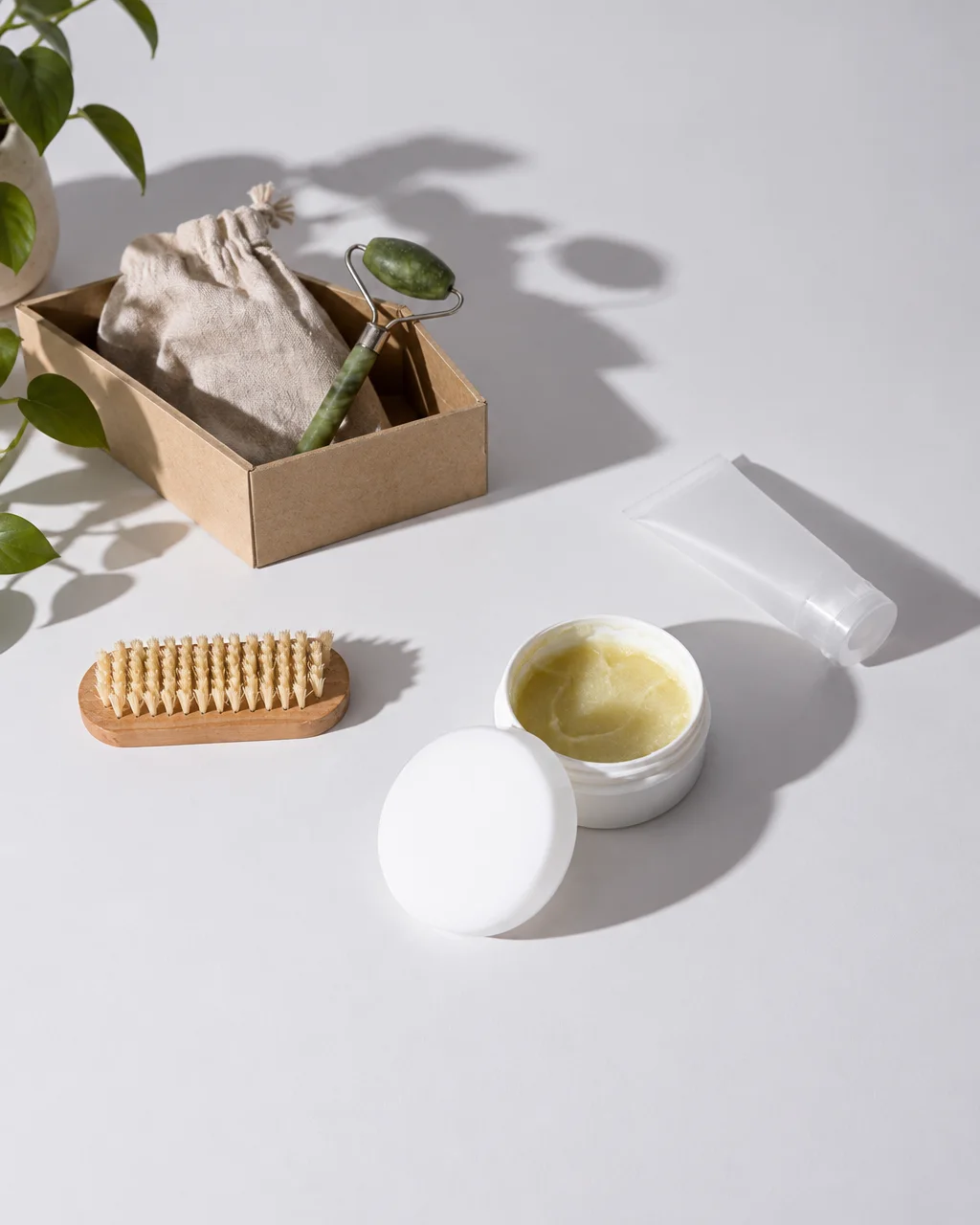 An open cosmetic balm jar sits beside a jade roller, wooden brush, and squeeze tube under bright natural light.