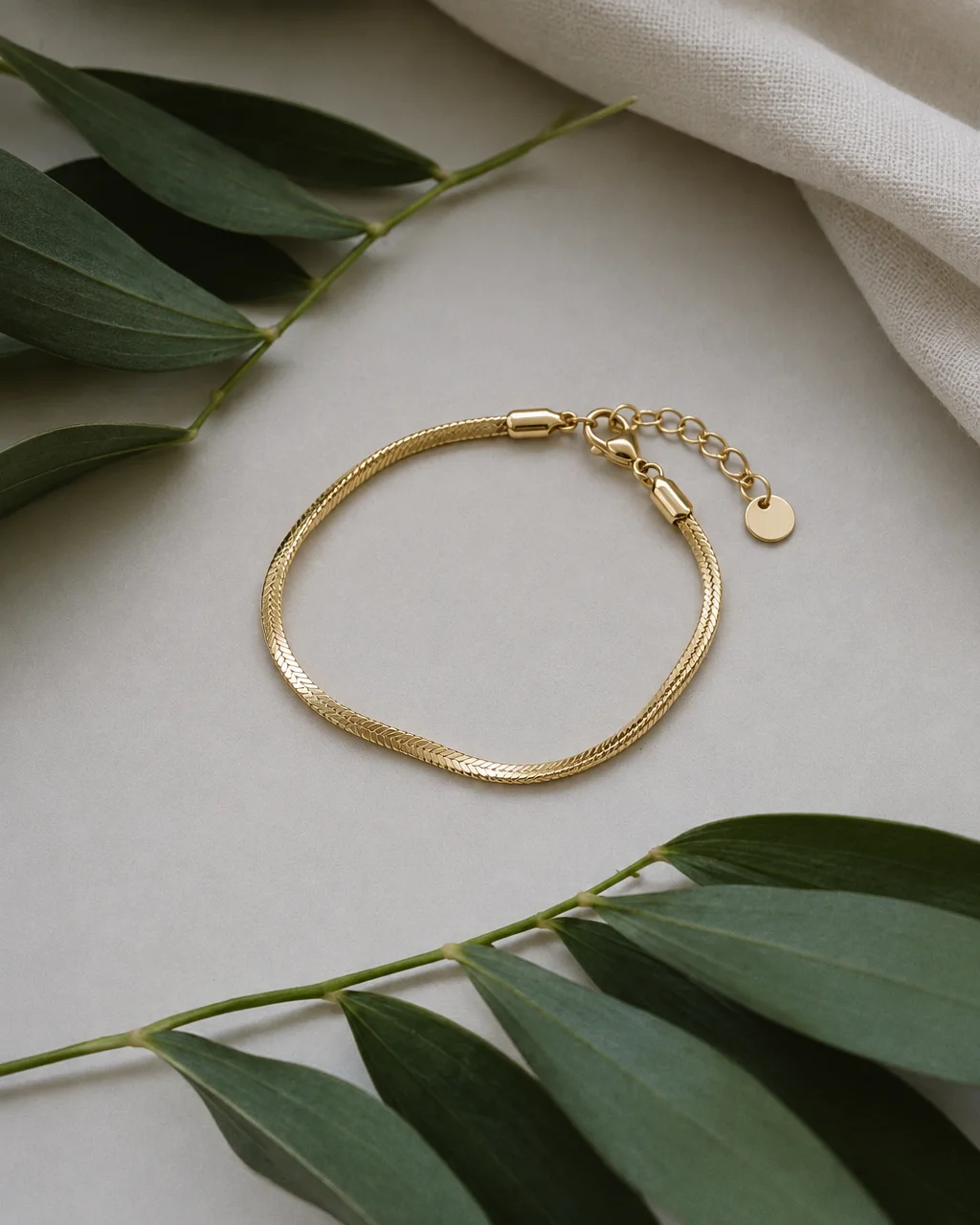 A gold herringbone bracelet laid flat on a pale surface between fresh green foliage and soft linen.