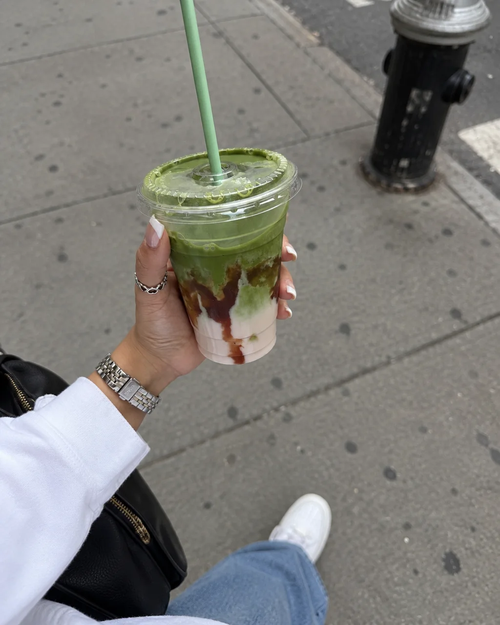 A person holding a layered iced matcha drink in a clear plastic cup while standing on a concrete city sidewalk.