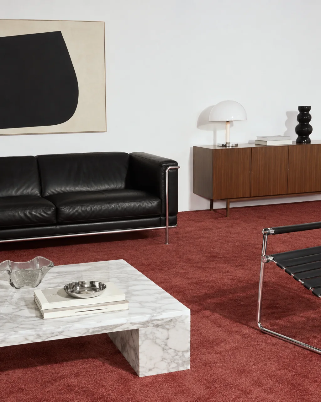 A modernist living space with a black leather sofa, low marble coffee table, and sideboard over red carpeting against white walls.
