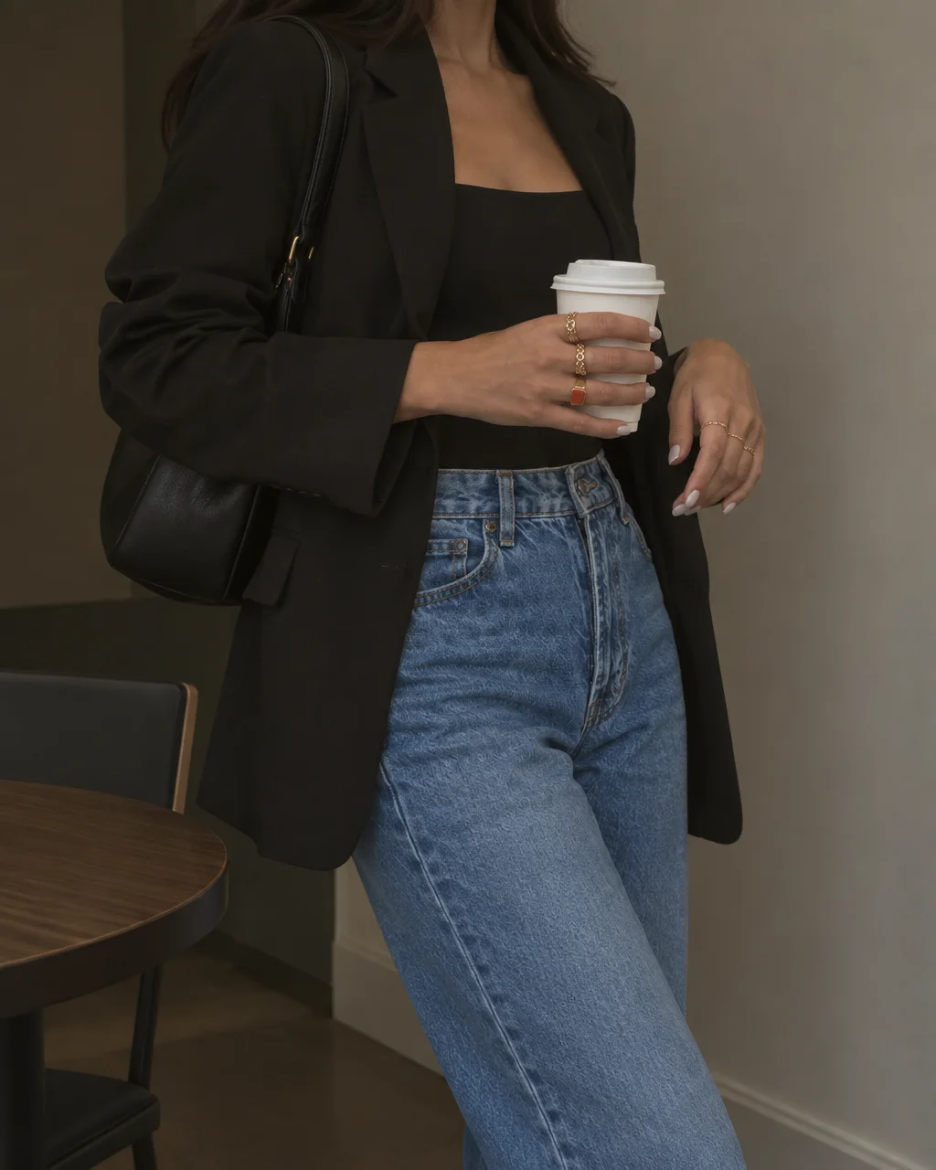 A woman wearing a tailored black blazer and blue denim jeans holding a takeaway coffee cup in a neutral interior.