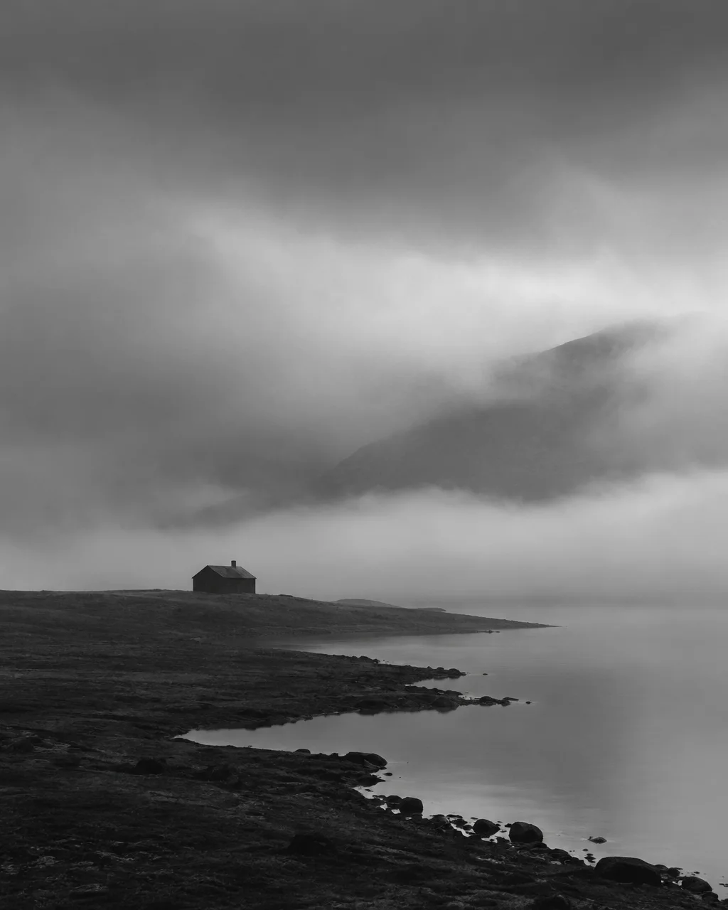 A solitary dark cabin stands on a rocky shoreline beneath dense fog rolling over a calm lake in black and white.