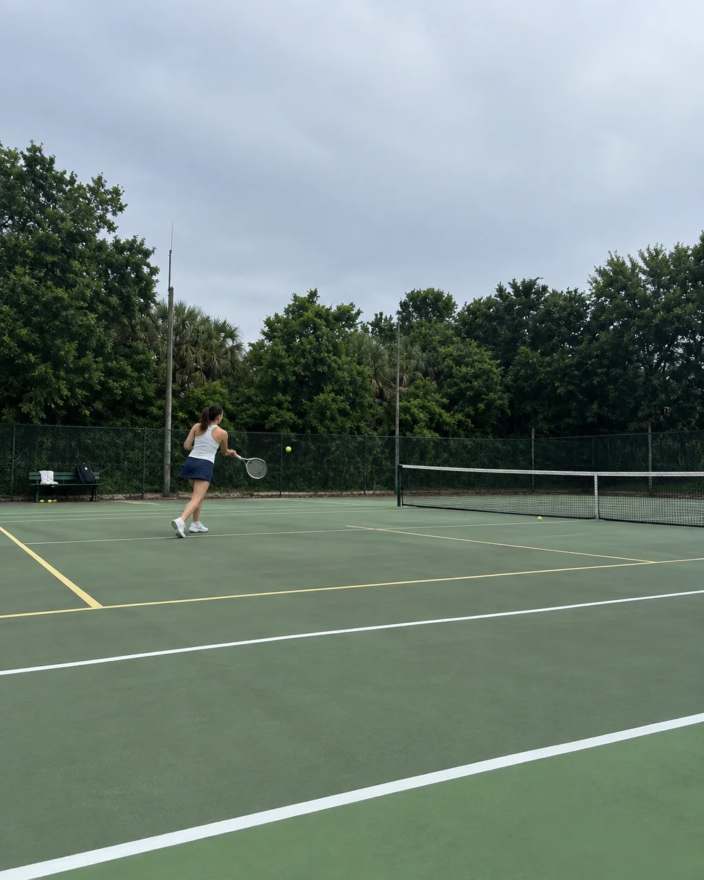 A woman swings her racket to return a tennis ball on an outdoor green court surrounded by lush trees under an overcast sky.