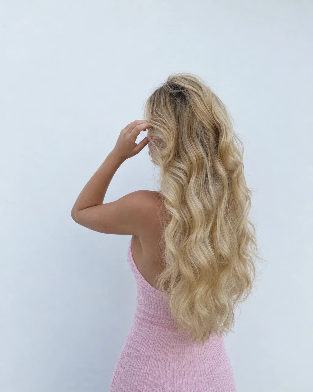 Back view of a woman with long wavy blonde hair wearing a pink textured dress against a plain white background.