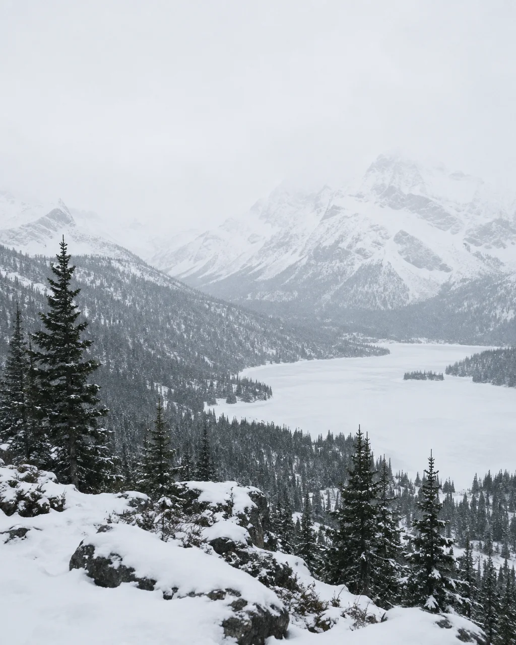 A high-angle view of a snow-covered mountain valley with a frozen lake and pine forests under an overcast sky.