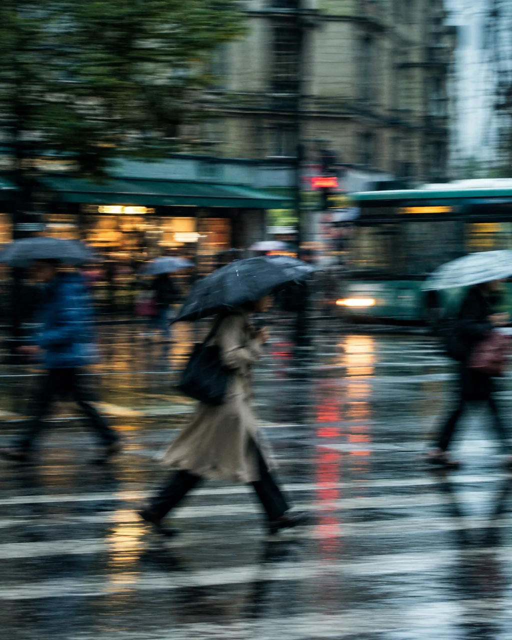 Motion-blurred pedestrians holding umbrellas walk across a wet city street with warm shop lights reflecting on the slick pavement.