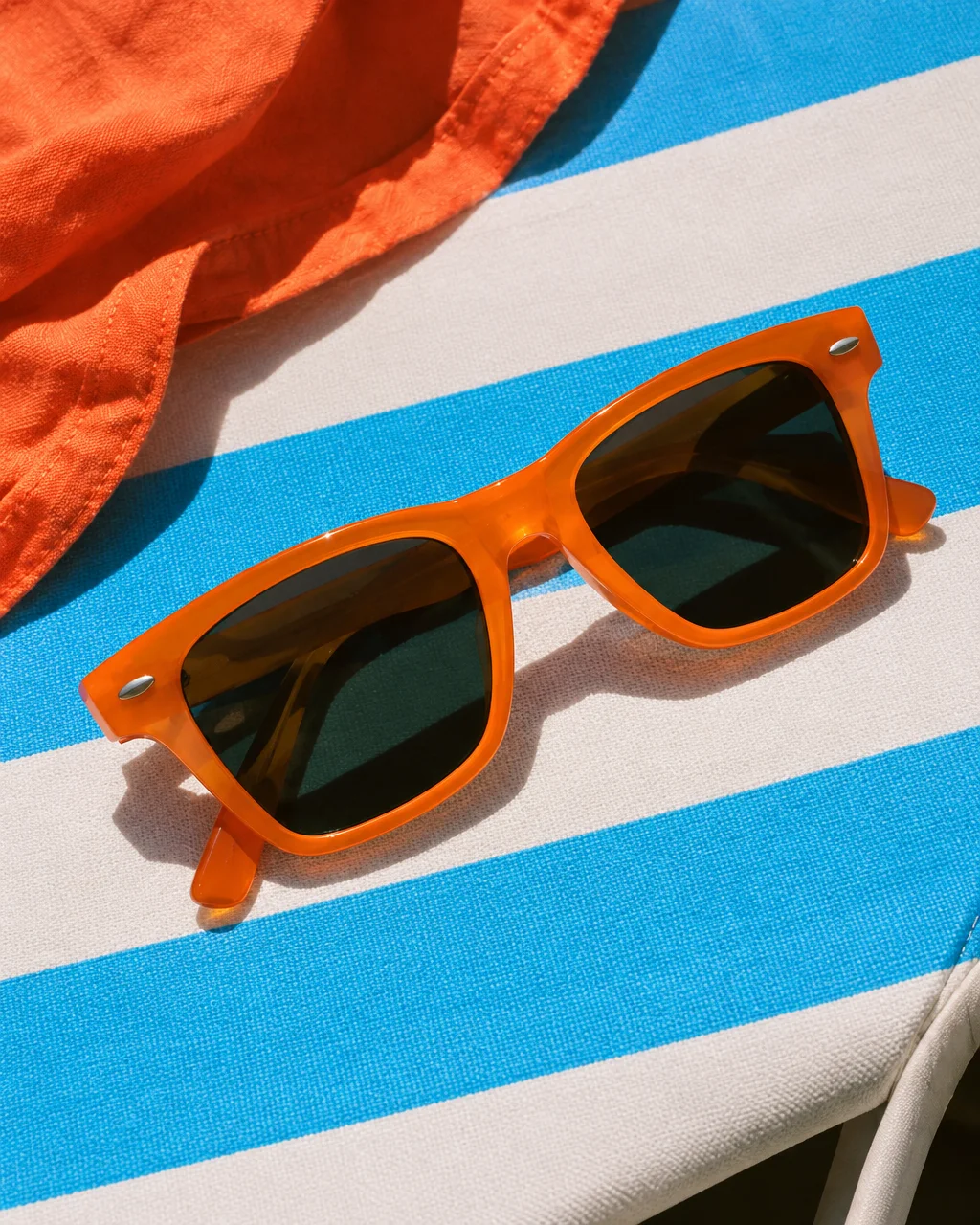 A pair of bright orange acetate sunglasses rests on a blue and white striped sun lounger beside vivid orange linen under strong direct sunlight.