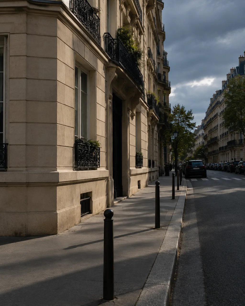 A quiet city sidewalk lined with black bollards alongside an ornate limestone apartment facade beneath an overcast sky.