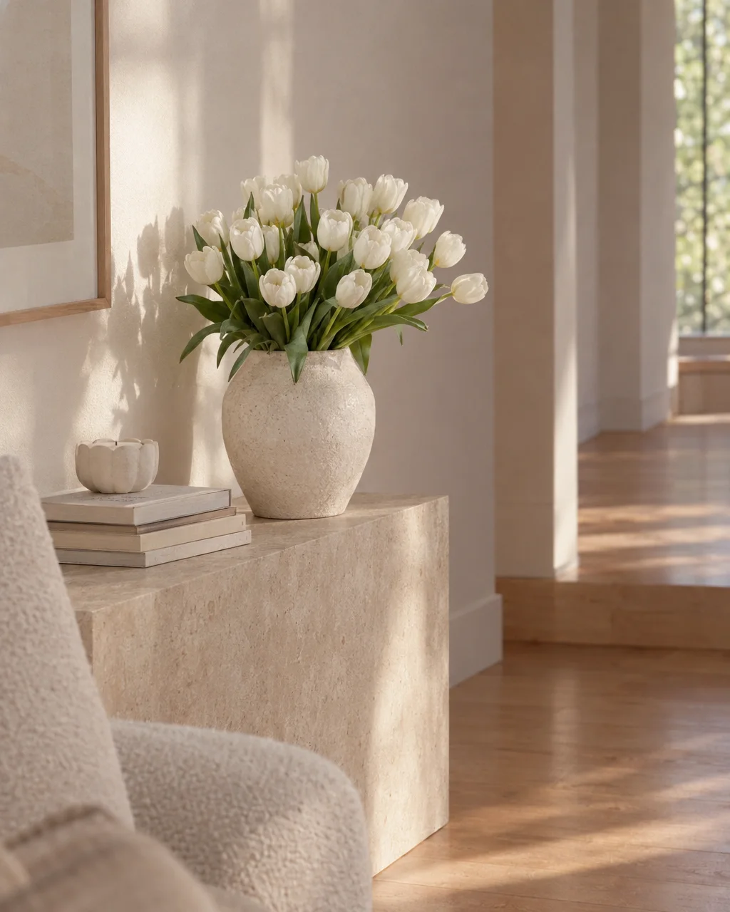 A bouquet of white tulips in a textured ceramic vase on a stone console inside a sunlit, minimalist neutral interior.
