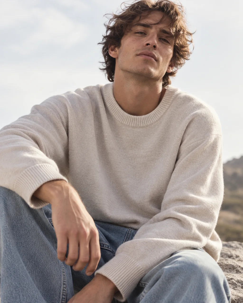 A young man wearing a relaxed cream knit crewneck sweater sits outdoors under a soft sky.
