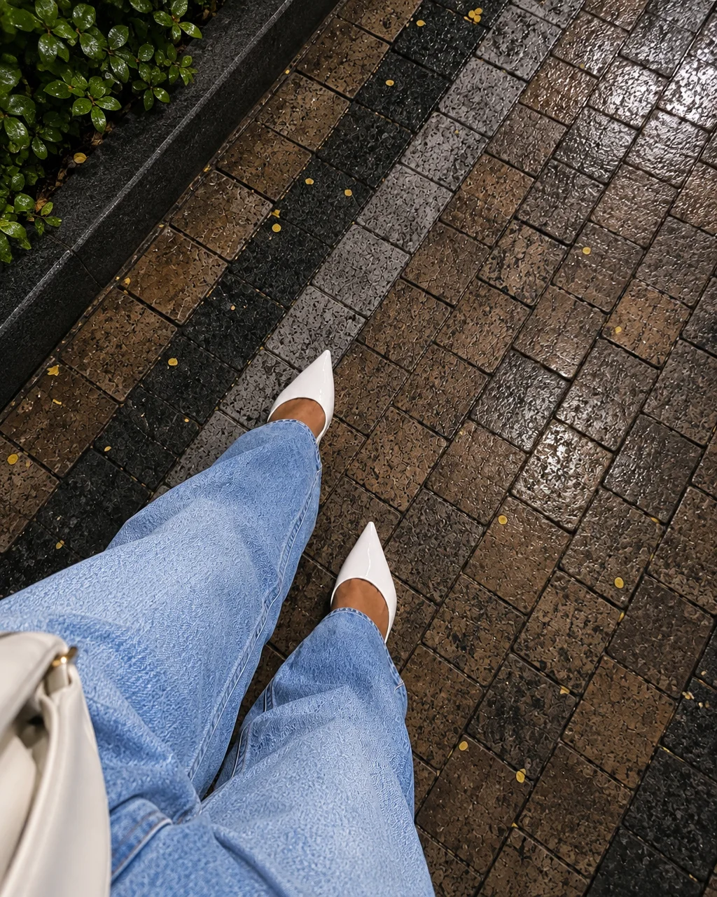 A downward perspective of pointed white pumps and light wash jeans stepping across wet tiled pavement beside green foliage.