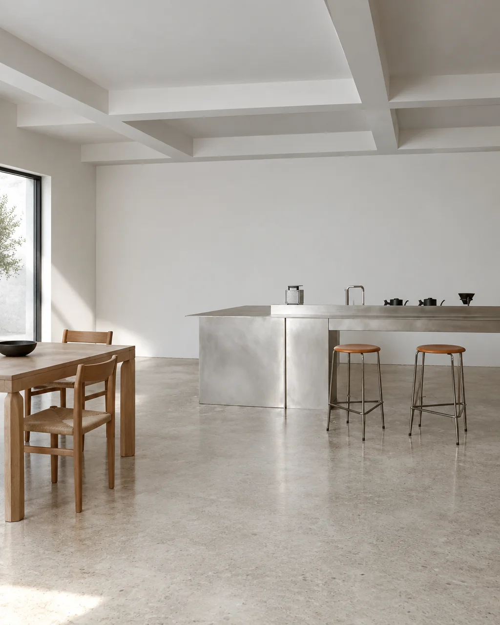 A minimalist open interior with a brushed steel kitchen island, bar stools, and a wooden dining table set on polished concrete.