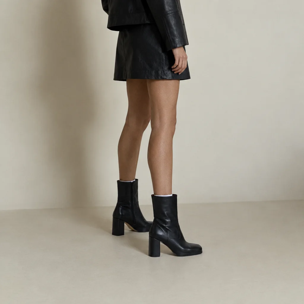 A model wearing a black leather miniskirt and heeled ankle boots stands against a muted cream studio backdrop.