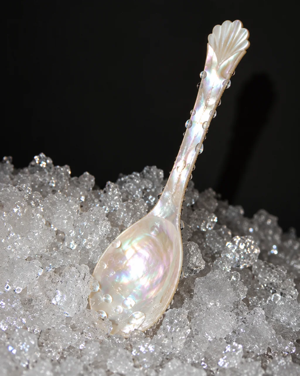 An iridescent mother-of-pearl spoon covered in water droplets rests in a mound of crushed ice against a dark backdrop.