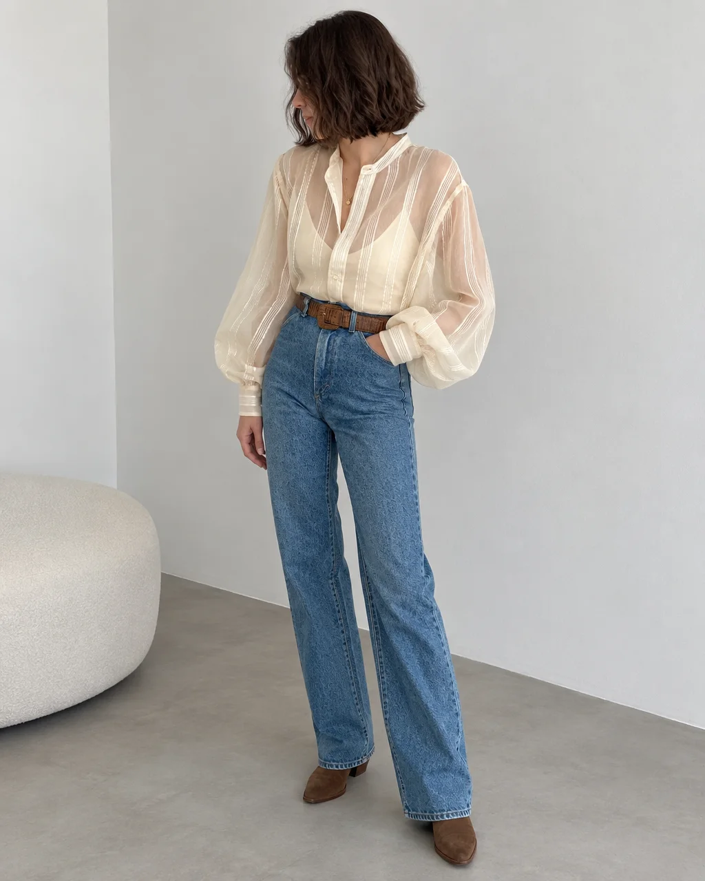 A woman wearing a sheer cream striped blouse tucked into high-waisted blue jeans stands in a minimalist interior space.