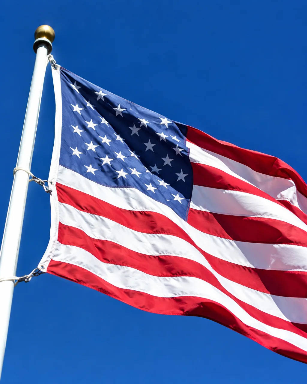 An American flag flutters from a white flagpole against a vivid, cloudless blue sky.