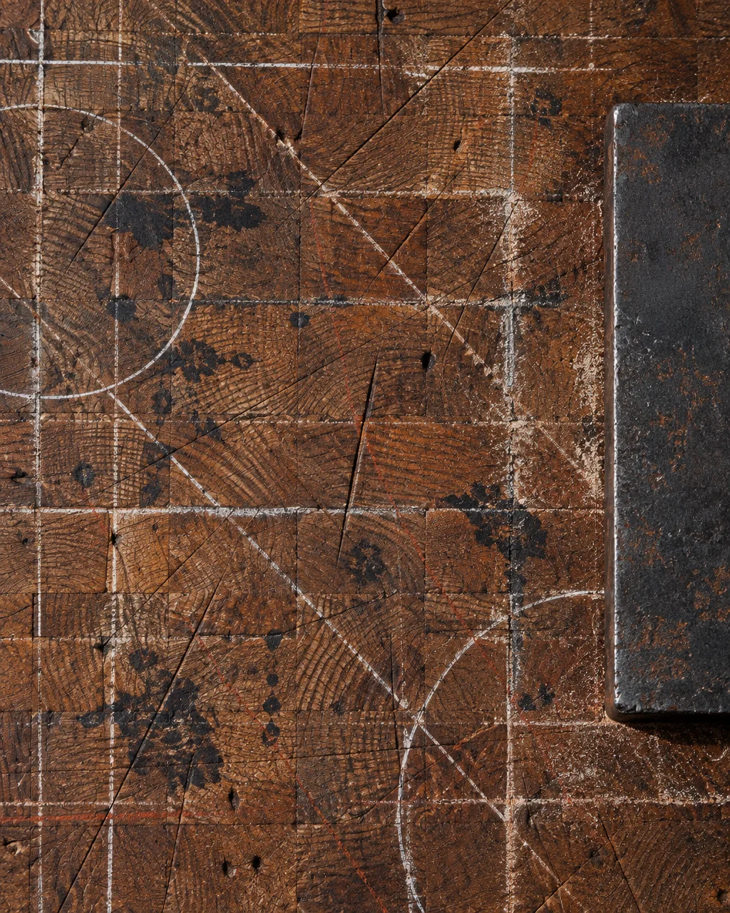 A heavily worn end-grain wooden workbench scored with white chalk gridlines, dark ink stains, and cutting marks next to a thick steel block.