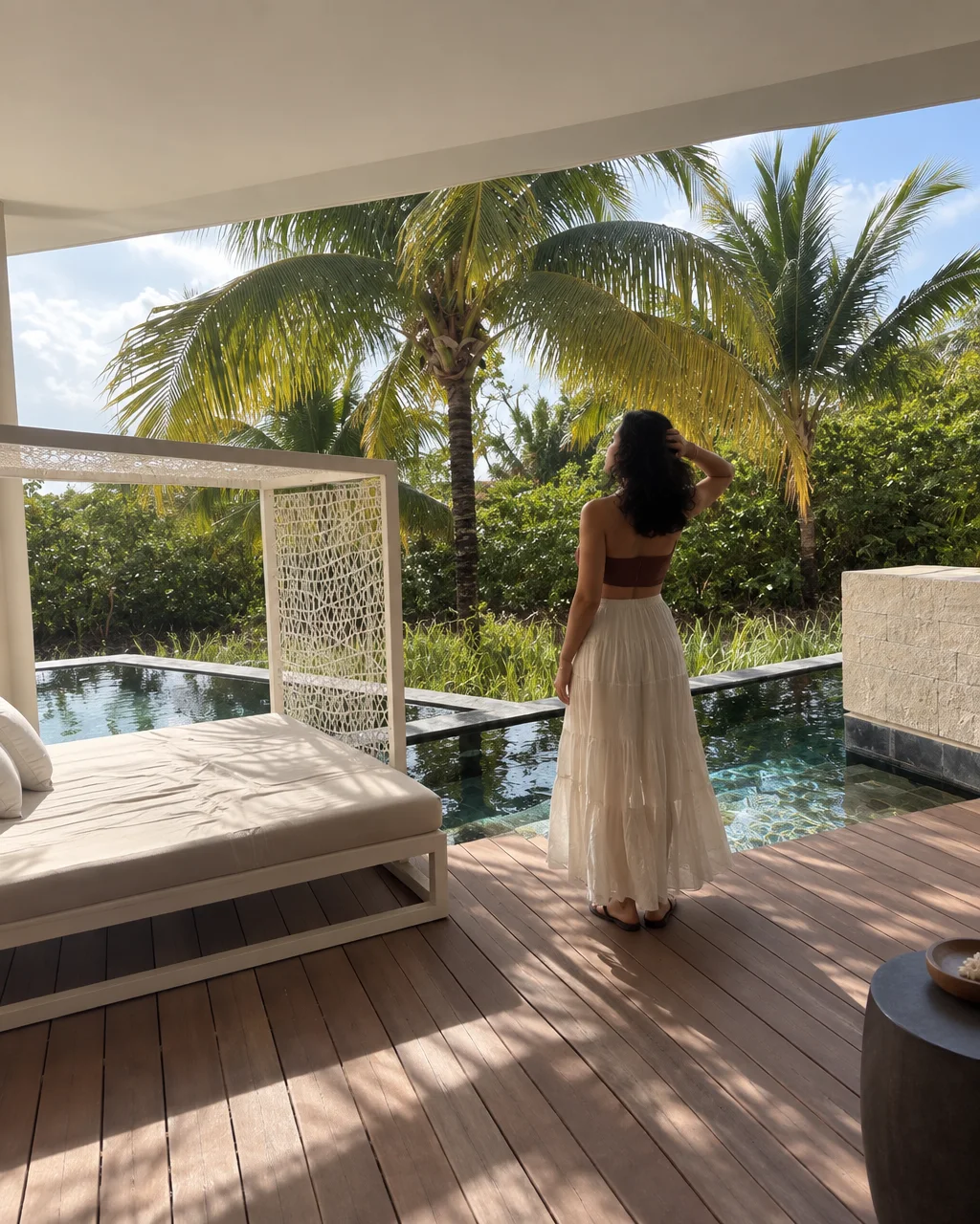 A woman in a white skirt stands on a wooden deck beside a private pool framed by lush tropical palm trees.