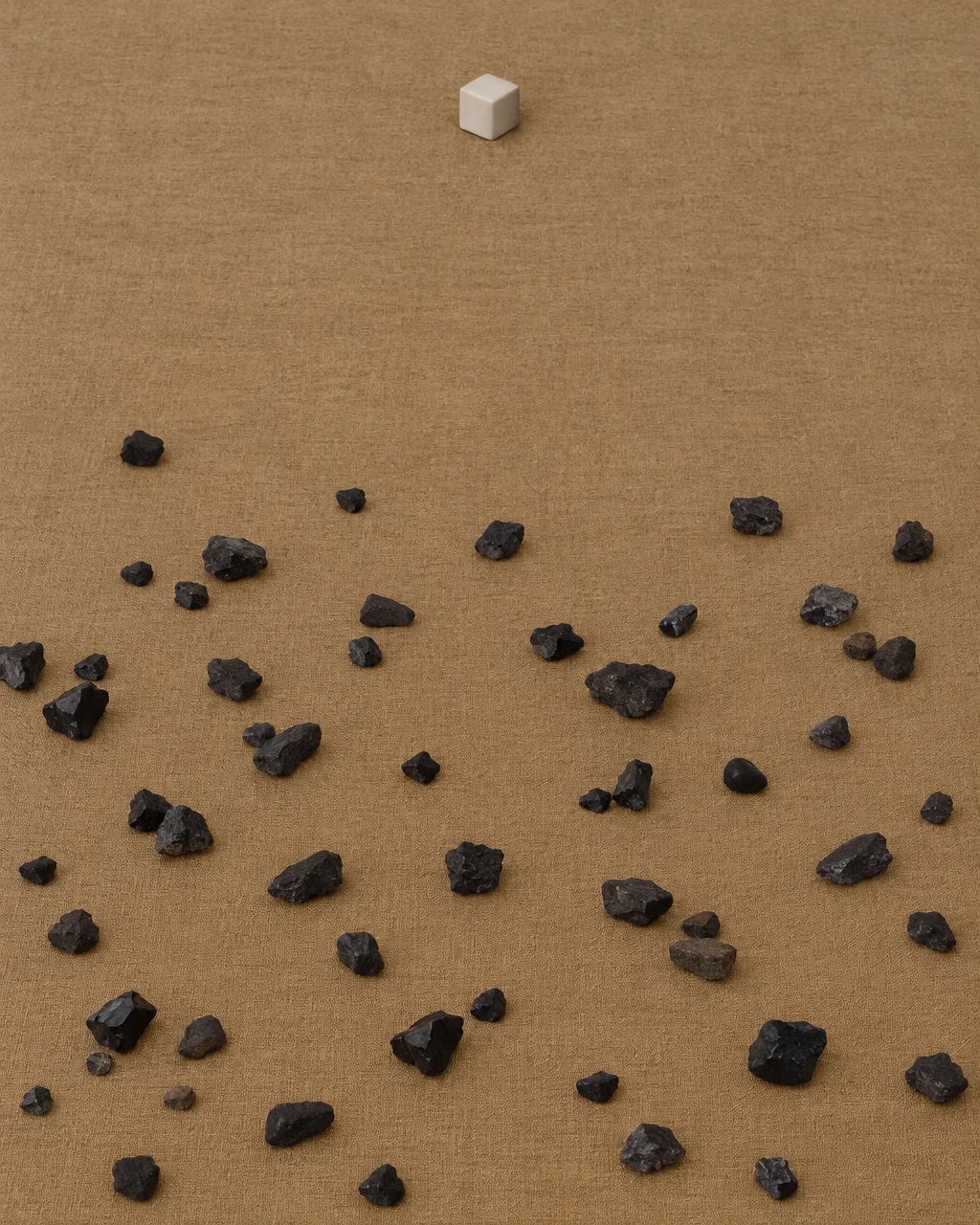 A single white cube rests alone above scattered rough black stones on a textured ochre canvas surface.