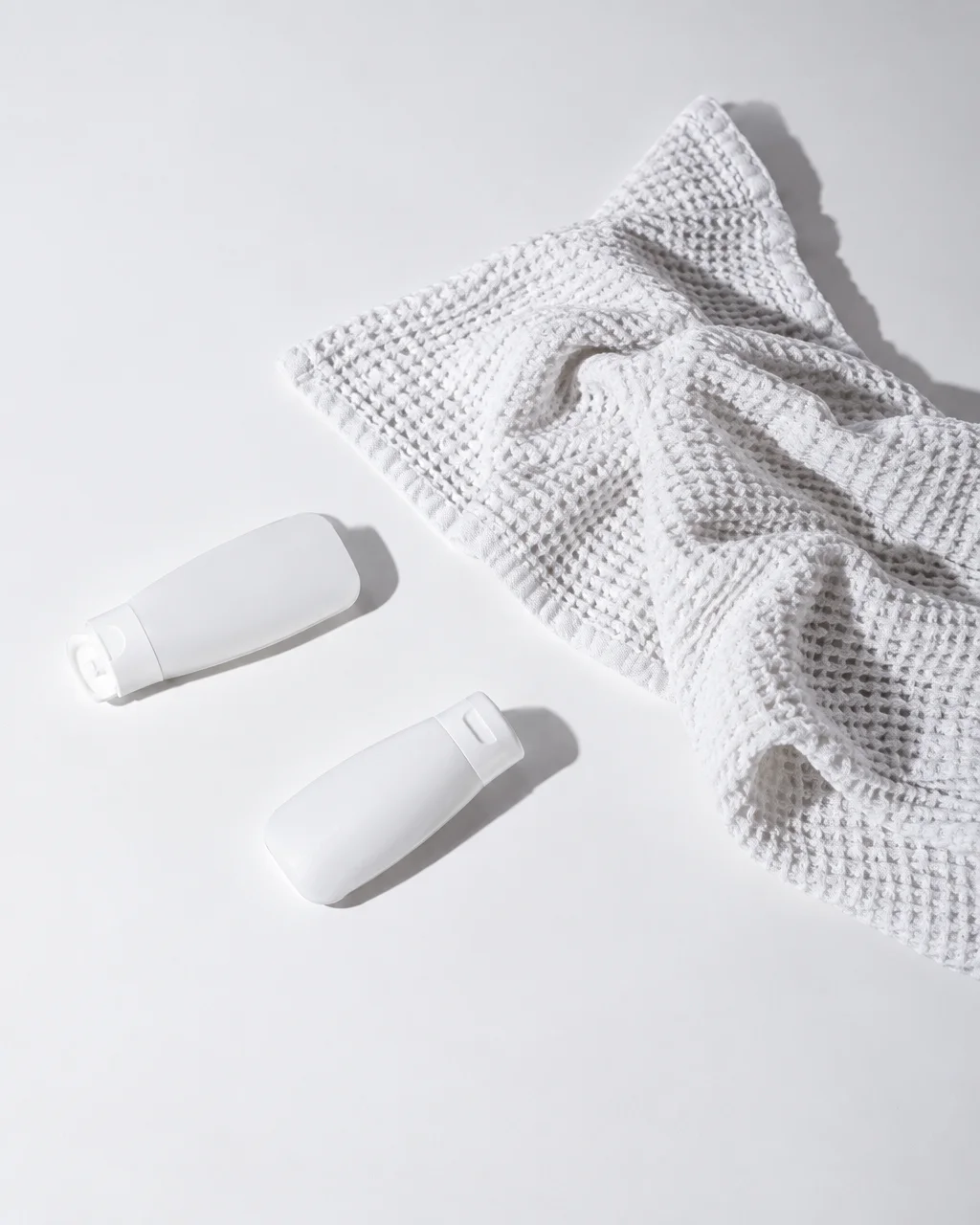 Two blank white cosmetic bottles lie beside a folded waffle-weave towel on a clean white studio surface under crisp light.