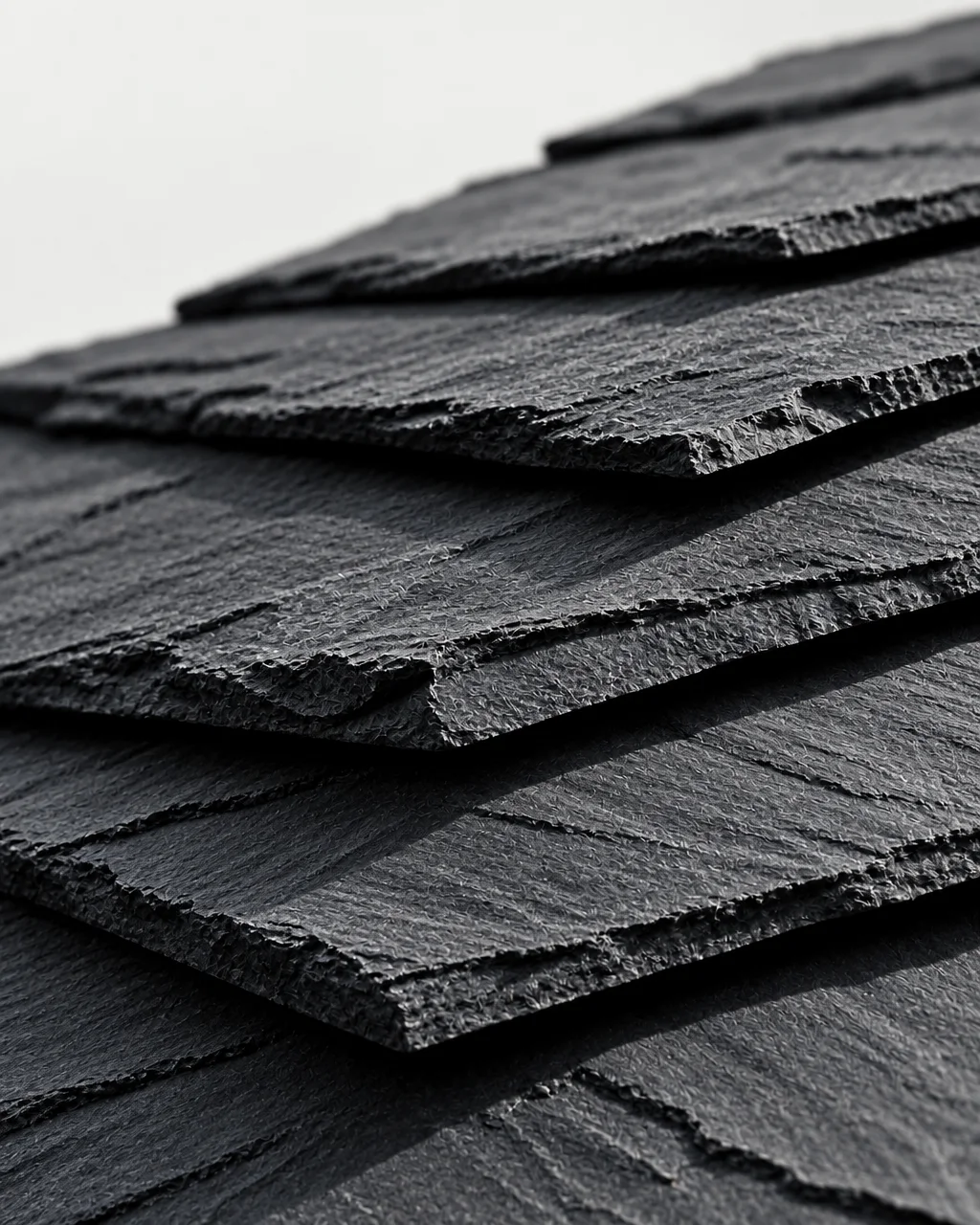 A close-up view of dark, rough-hewn slate roof shingles with textured edges casting sharp diagonal shadows.