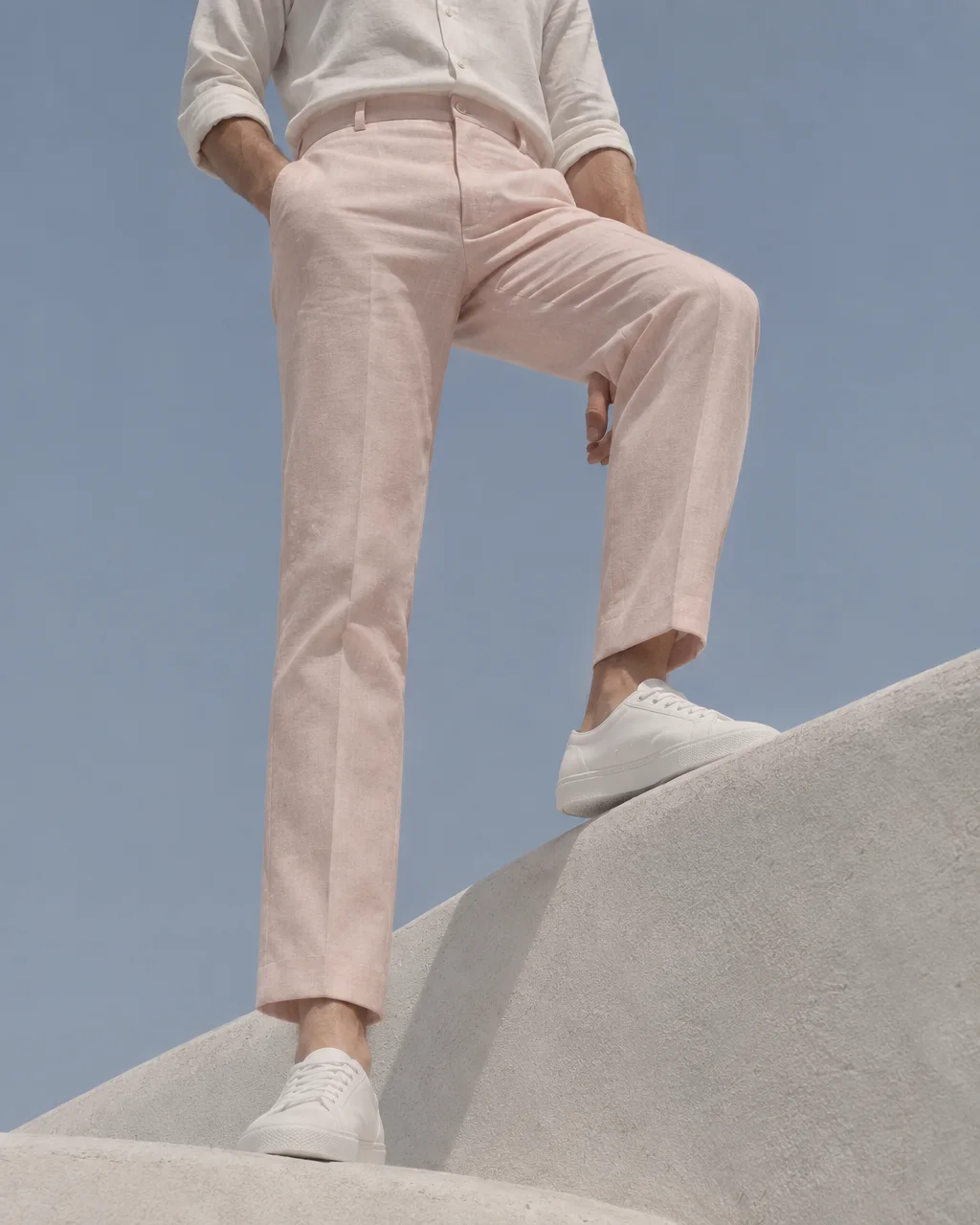 A low-angle view of a person standing on white concrete steps in pale pink linen trousers and white sneakers against a clear sky.