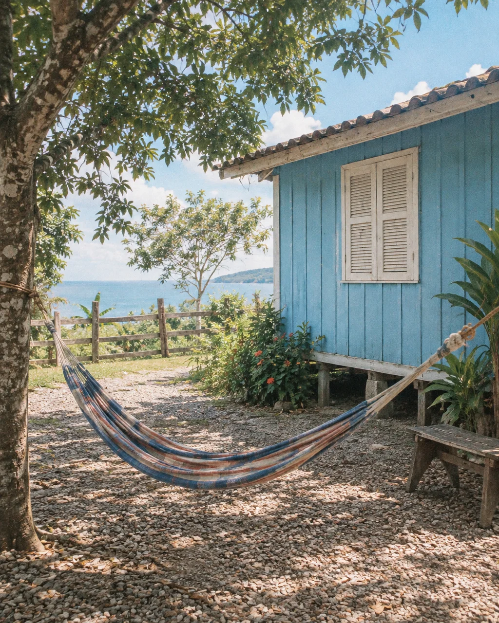 A striped hammock suspended between a tree and a rustic blue wooden cabin overlooking the ocean on a sunny day.