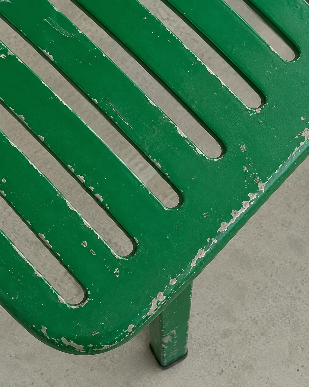 A close-up view of a weathered green metal slatted chair with peeling paint over a neutral concrete surface.