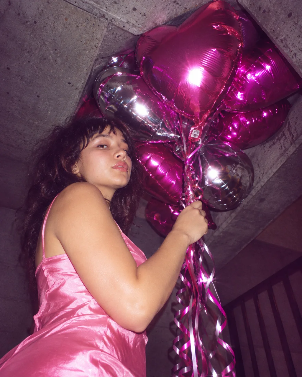 A young woman in a pink satin slip dress holds a cluster of shiny metallic heart balloons beneath a raw concrete staircase.