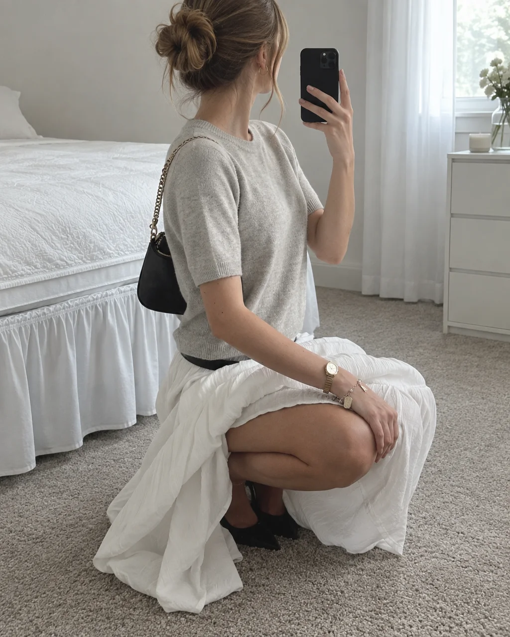 A woman wearing a grey knit top and a flowing white maxi skirt crouches in a bright bedroom to take a mirror selfie.