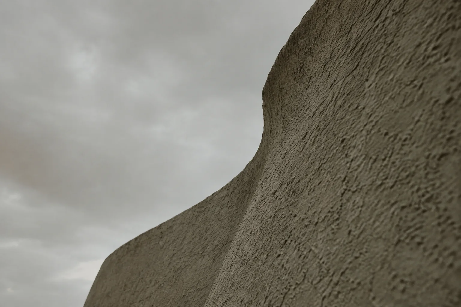 A textured, curving adobe wall slopes upward against a muted, overcast sky.