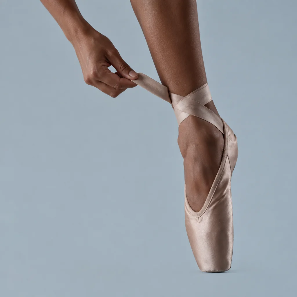 A ballerina adjusts the satin ribbon of a silk pointe shoe against a muted blue studio backdrop.