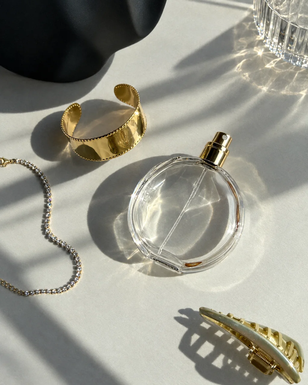 A round glass perfume bottle, gold cuff bracelet, and accessories arranged under direct striped sunlight with soft caustic reflections.