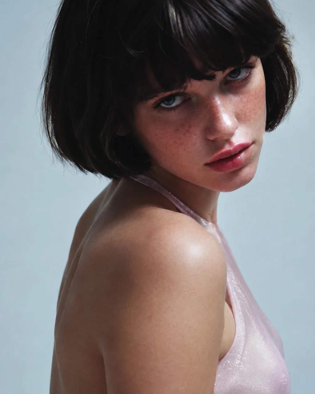 A close studio portrait of a woman with a dark bob haircut and freckles looking over her bare shoulder against a cool grey backdrop.