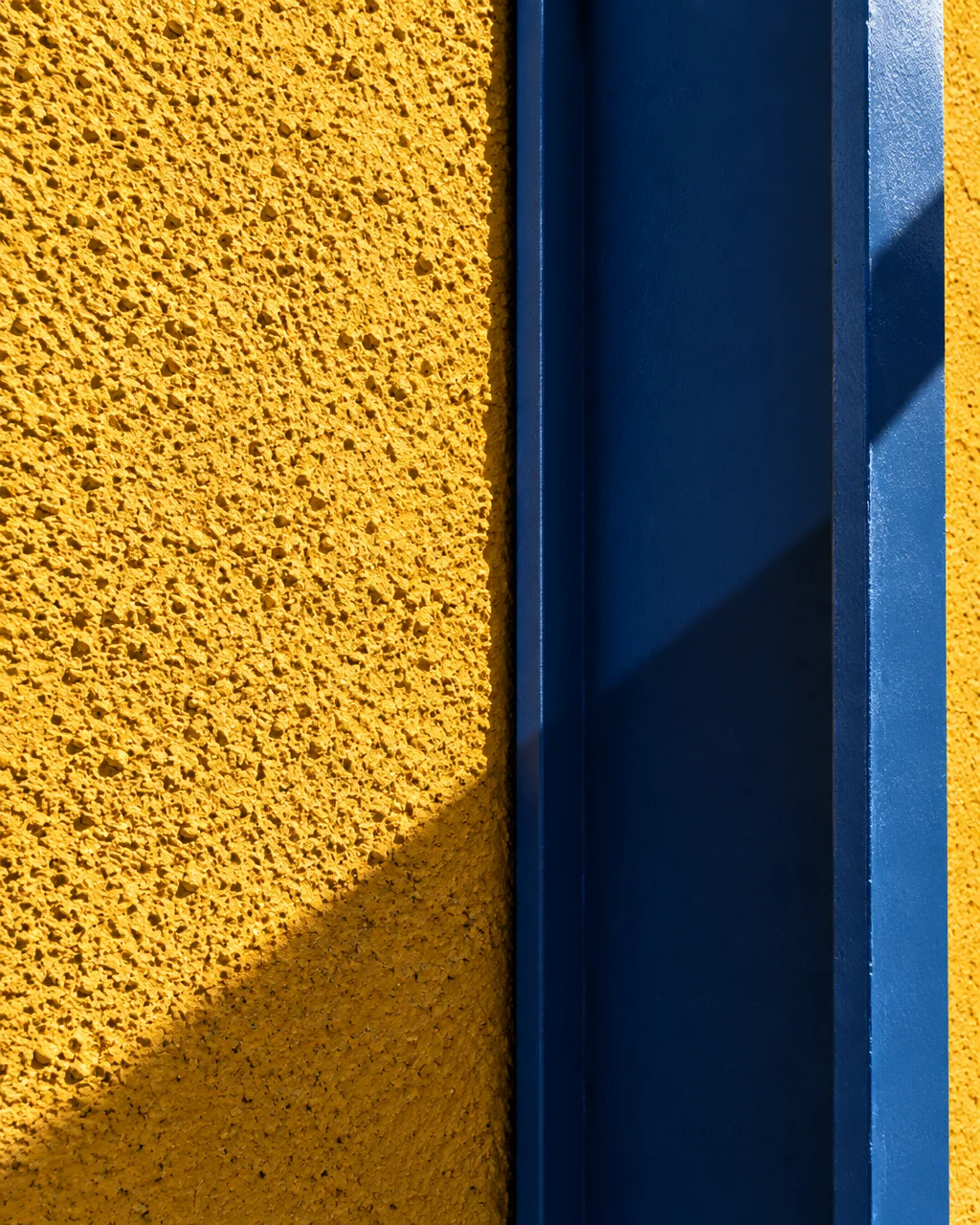 A close-up architectural detail of a vibrant textured yellow stucco wall paired with a bold blue painted steel beam under direct sunlight.