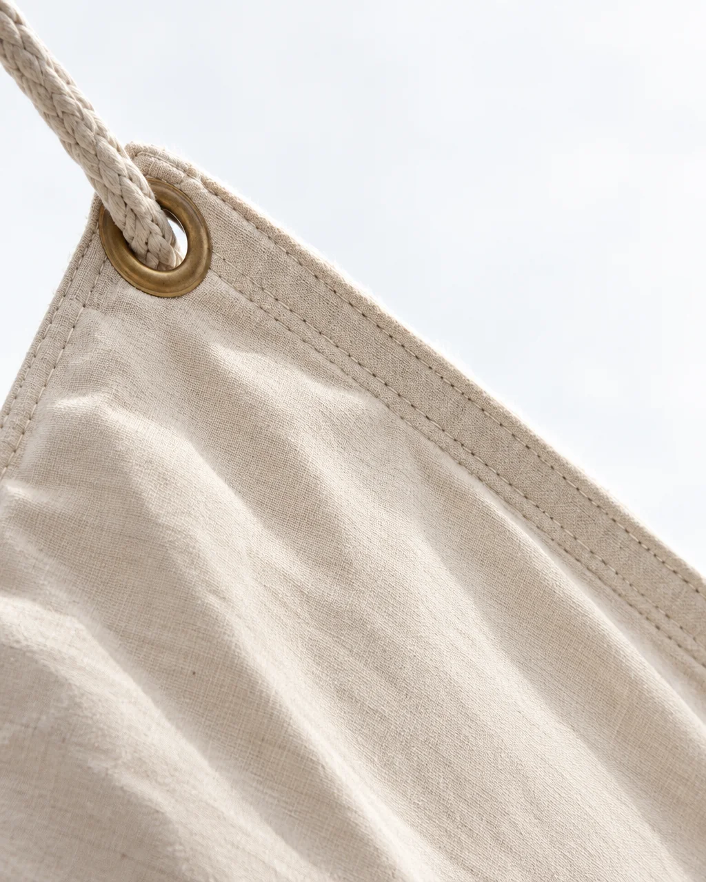 A detailed view of a cream linen canvas canopy corner with heavy stitching, a brass grommet, and braided rope against bright daylight.