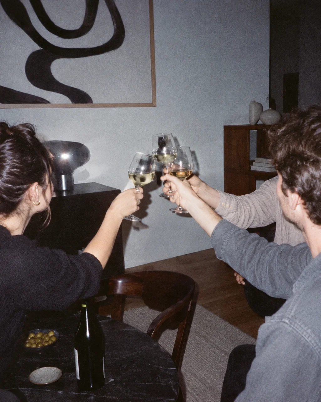 A group of friends toasting with glasses of white wine around a dark table in a stylish, dimly lit apartment.
