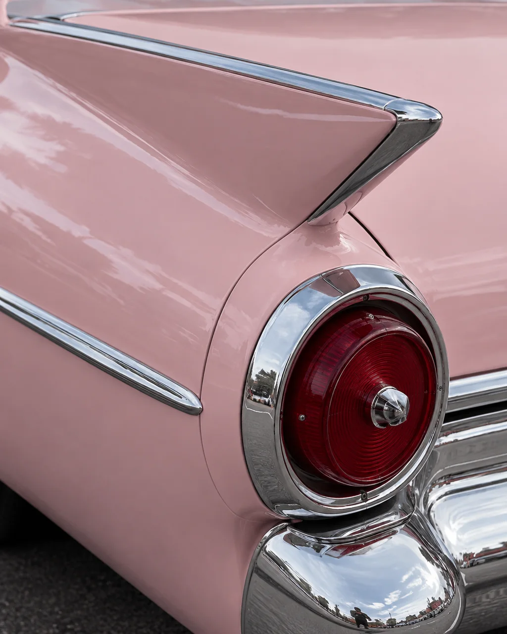 A close-up view of a pastel pink vintage car showcasing its sculpted tailfin, polished chrome bumper, and round red taillight.